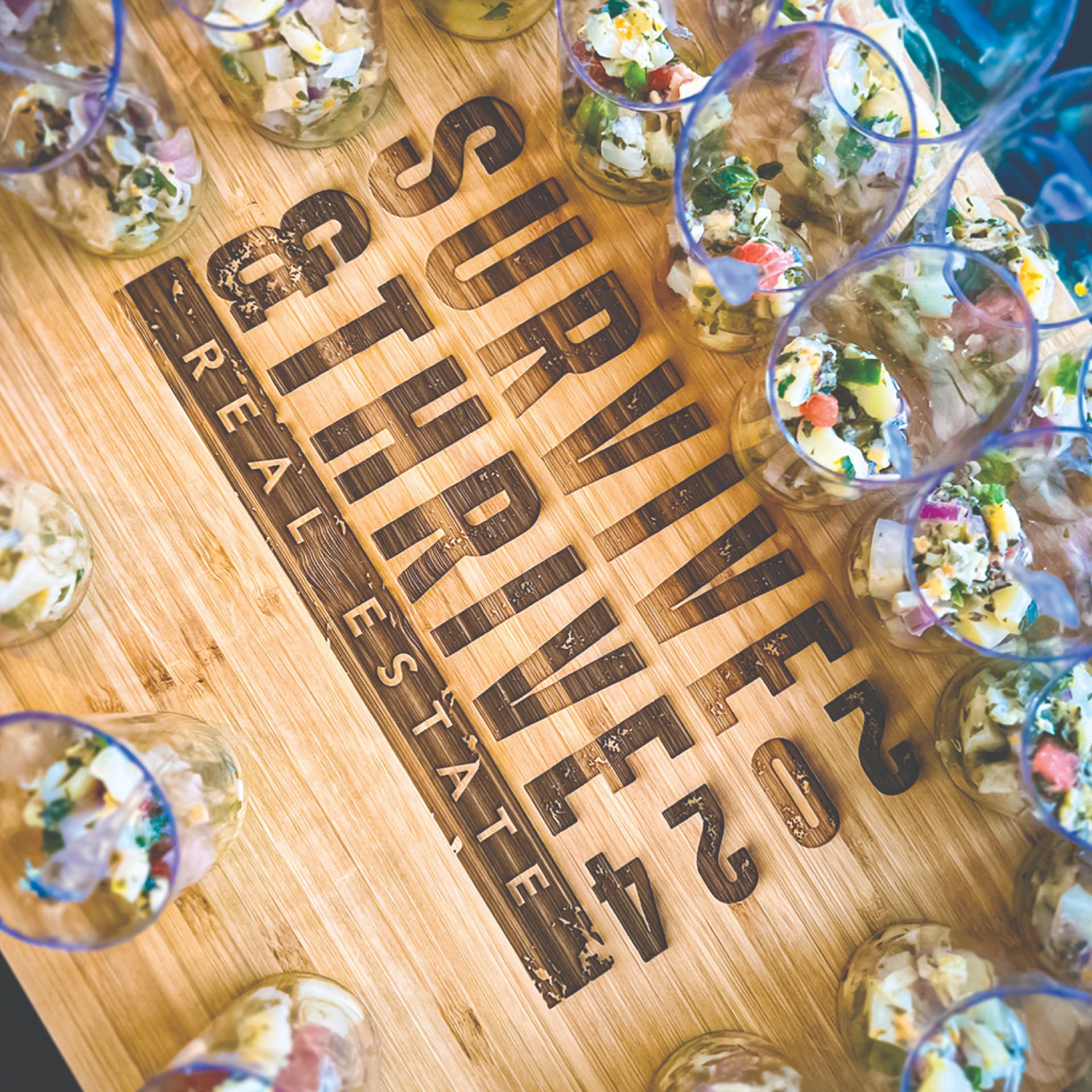 Laser-engraved bamboo serving board branded Survive & Thrive 2024 Real Estate, set with individually plated ceviche cups at an event